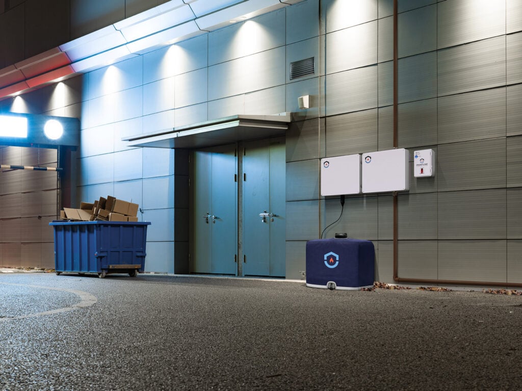 A photo showing Frontline's commercial wildfire defense system mounted to a building exterior wall near a back entrance and dumpster.