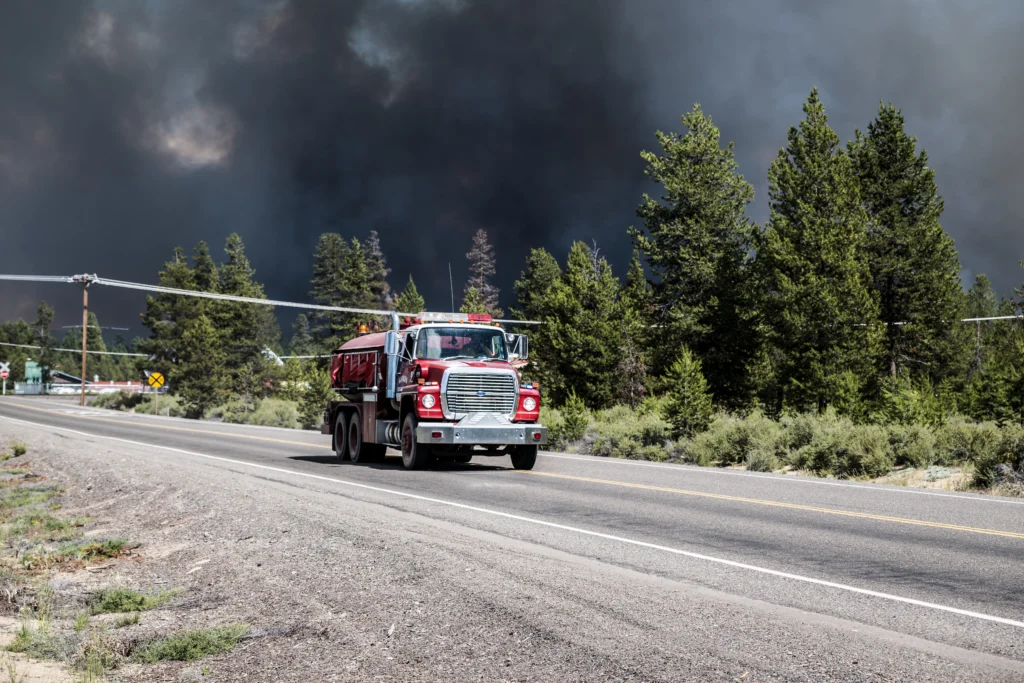 A red firefighting truck drives down a highway ahead of a massive wall of dark smoke, showing emergency response and getting help during forest fires in Oregon.
