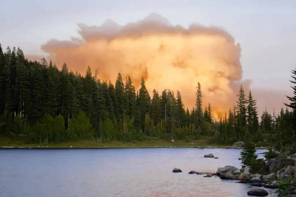 A large plume of glowing orange smoke rises behind a dense evergreen forest and a mountain lake during the peak of the wildfire season in Oregon.