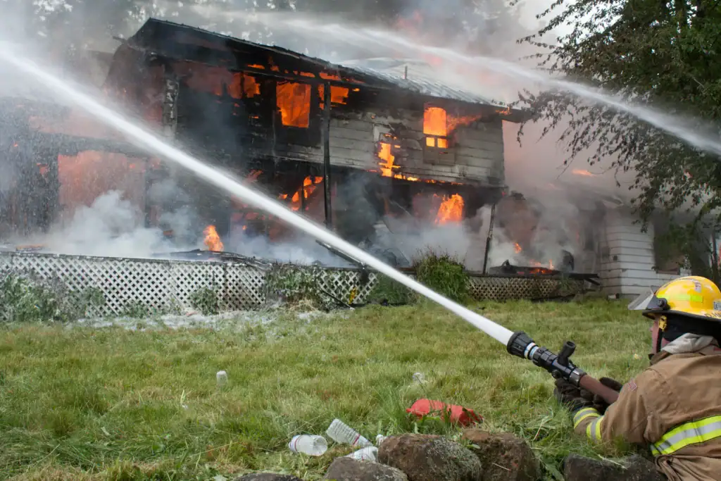 A firefighter spraying a hose on a burning structure, illustrating high-volume suppression tactics compared to precision wildfire prevention.