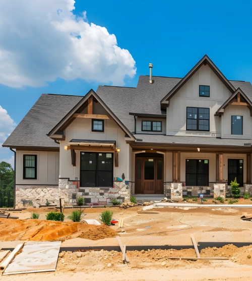 Large multi-story modern house under construction on a bare dirt lot under a sunny sky.