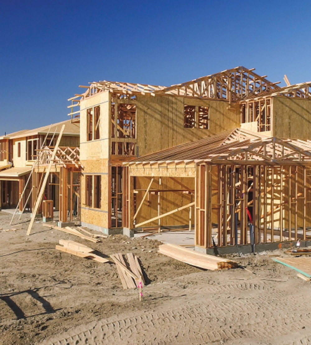 Wooden frame of a multi-story home undergoing active reconstruction on a dirt lot under a blue sky.