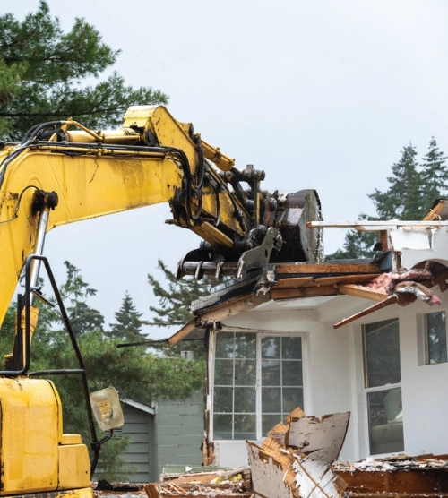 Yellow heavy excavator demolishing damaged roof structure and clearing debris from a home site.