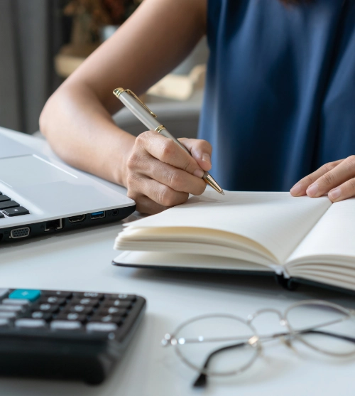 Person sitting at a desk writing in an open notebook beside a laptop, calculator, and eyeglasses.